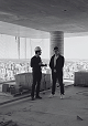 Dois profissionais conversam em pavimento em obra, com a cidade ao fundo.